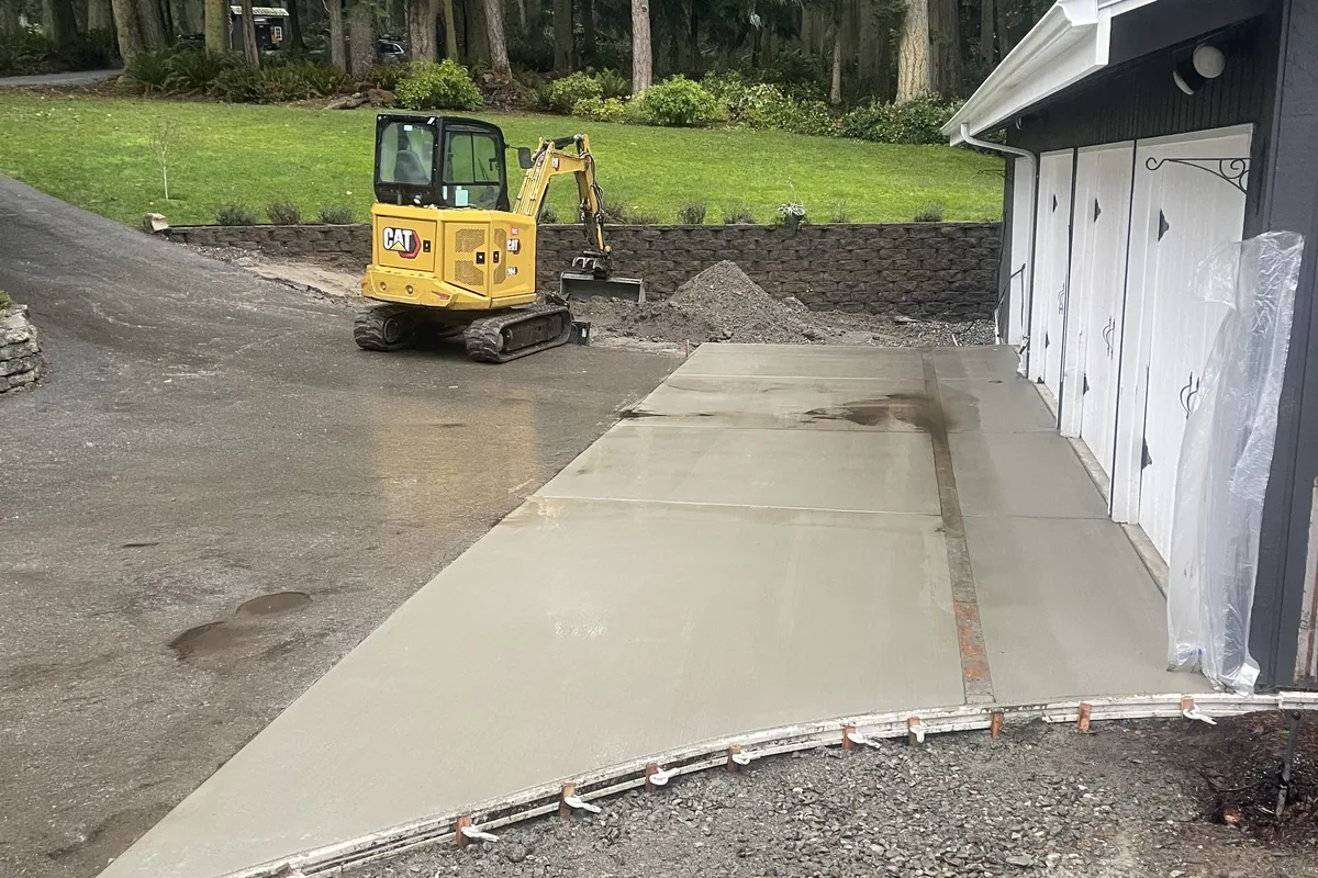 Mini excavator parked beside a freshly poured concrete apron in front of a garage, forms and stakes still on the front edge