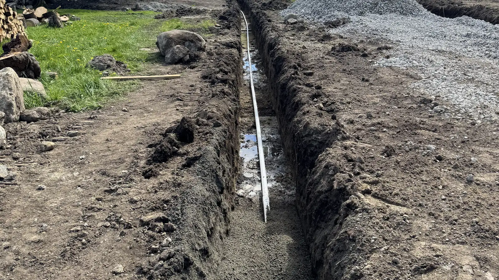 Long open trench with a white pipe along its wet bottom, between a lawn and a pile of crushed rock
