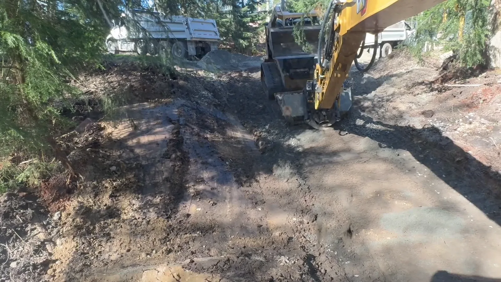 Excavator working a cleared strip of wet ground in the woods, with dump trucks parked behind it