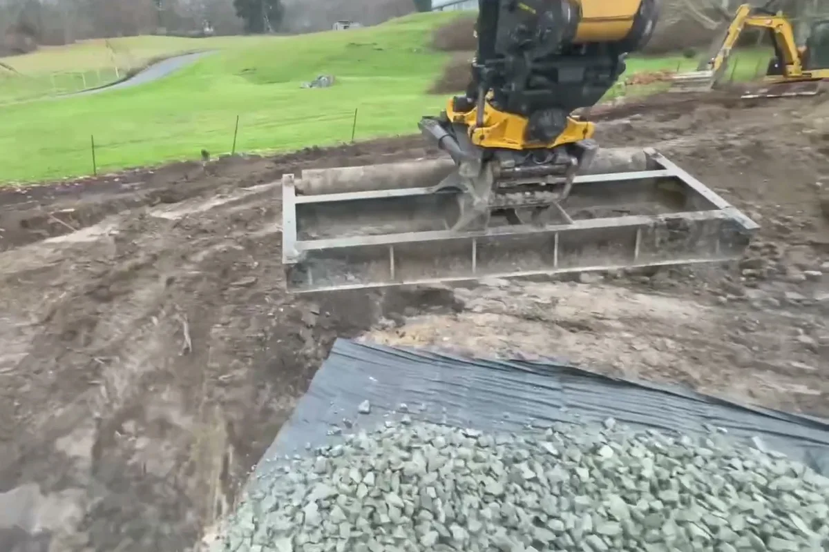 Tilt rotator grading attachment leveling base rock on a new gravel driveway beside a pasture